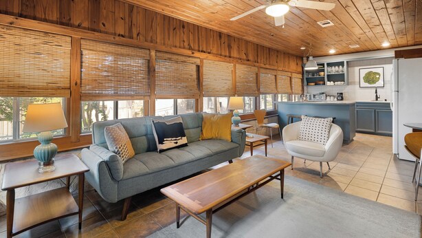 A cozy living room with a sofa, coffee table, and chairs, a kitchen area with cabinets and a refrigerator, and large windows with blinds. - Waterlily Serenity | 2 Bedroom | Creekside (Wimberley)