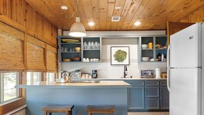 A kitchen with wooden ceiling, a large island with stools, and a refrigerator. - Waterlily Serenity | 2 Bedroom | Creekside (Wimberley)
