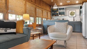 A cozy living room with a sofa, armchair, and table, a kitchen area with shelves and a refrigerator, and large windows with blinds. - Waterlily Serenity | 2 Bedroom | Creekside (Wimberley)