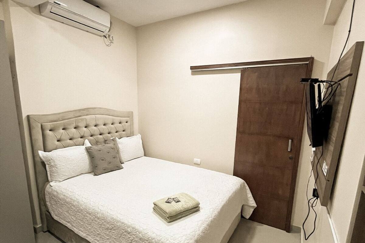 A bedroom with a bed, a TV mounted on the wall, and an air conditioning unit on the ceiling.