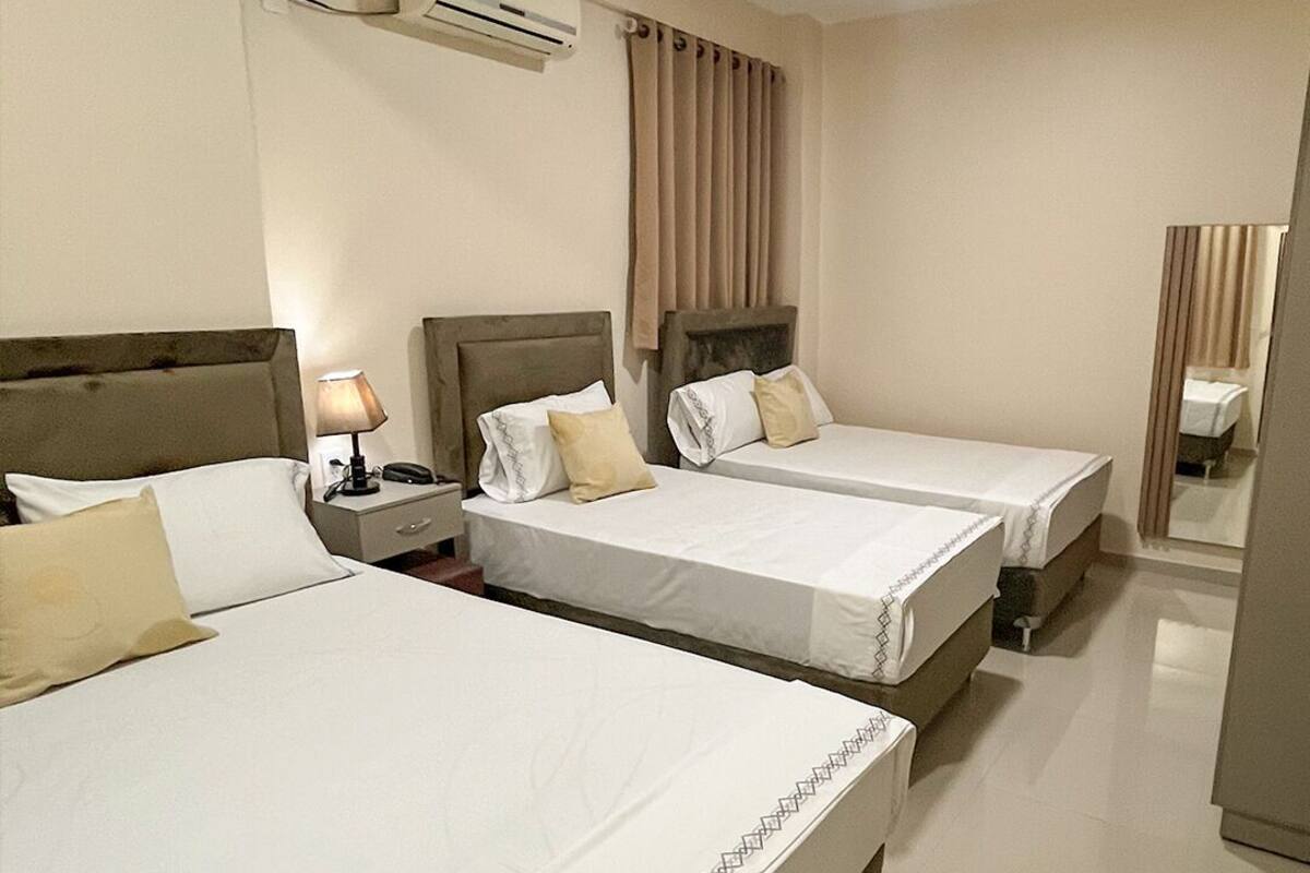 A hotel room with three single beds, a bedside table with a lamp, an air conditioning unit, and a mirror.