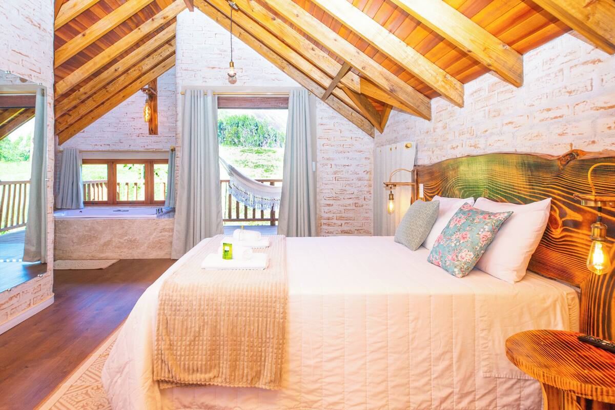 Une chambre avec un grand lit, des poutres apparentes en bois au plafond, une vue sur l’extérieur et un balcon.