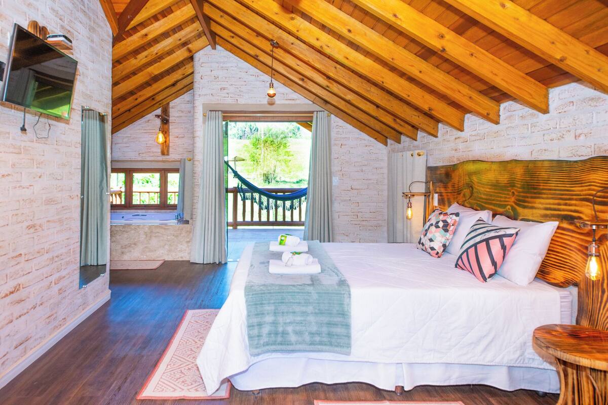 Une chambre avec un plafond en bois, un lit avec des oreillers, une table de chevet et une vue sur un hamac à l’extérieur.