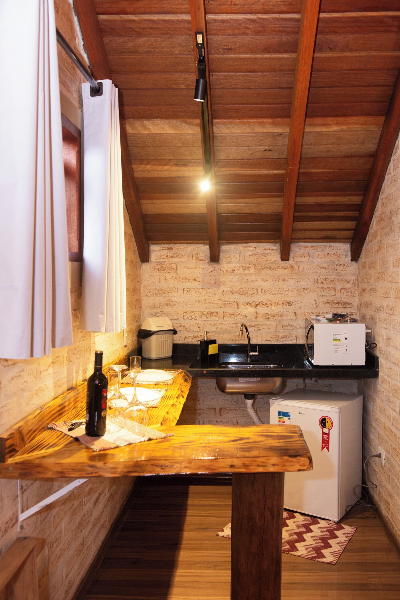 A compact kitchen with a wooden table, a sink, a refrigerator, and a brick wall.