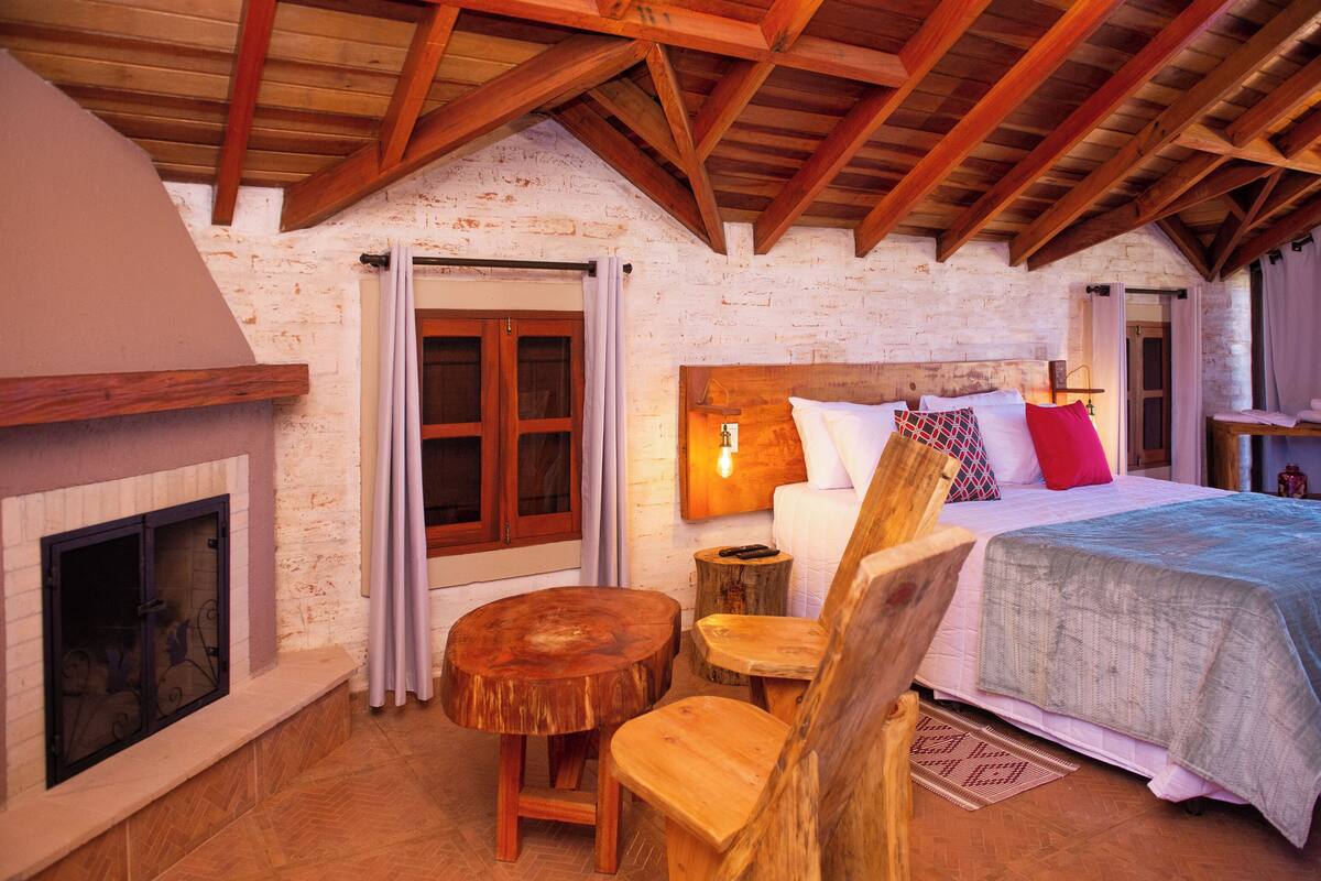Une chambre confortable avec un plafond en bois, une cheminée, un lit avec des oreillers et une petite table en bois.