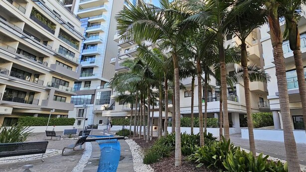 A multi-story residential building with balconies, palm trees, and a blue water feature.