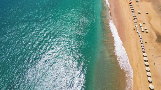 An aerial view of a beach with clear turquoise waters, a sandy shore, and a row of beach umbrellas. - Casa Pé na Areia (Fernando de Noronha)