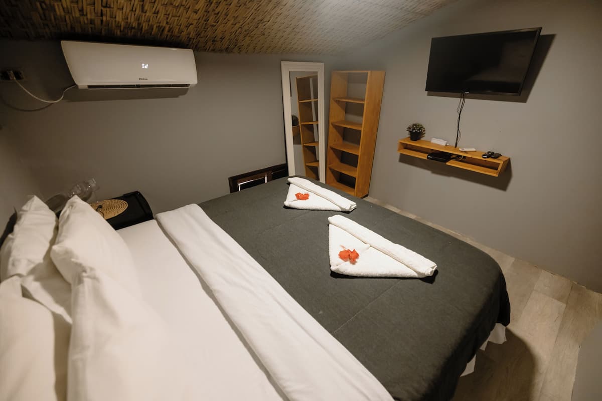 Una cama bien hecha con ropa de cama y toallas blancas, un televisor montado en la pared, un estante de madera con una planta y un armario con espejo.