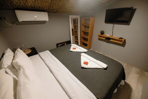 A neatly made bed with white linens and towels, a wall-mounted TV, a wooden shelf with a plant, and a wardrobe with a mirror.