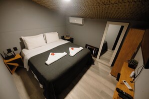 A hotel room with a large bed, a bedside table with a lamp, a wall-mounted air conditioner, and a wooden desk with a plant.