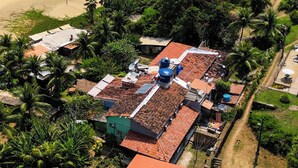 An aerial view of a residential area with multiple buildings, including a house with a red roof and a green building with a satellite dish. - Casa Pé na Areia (Fernando de Noronha)