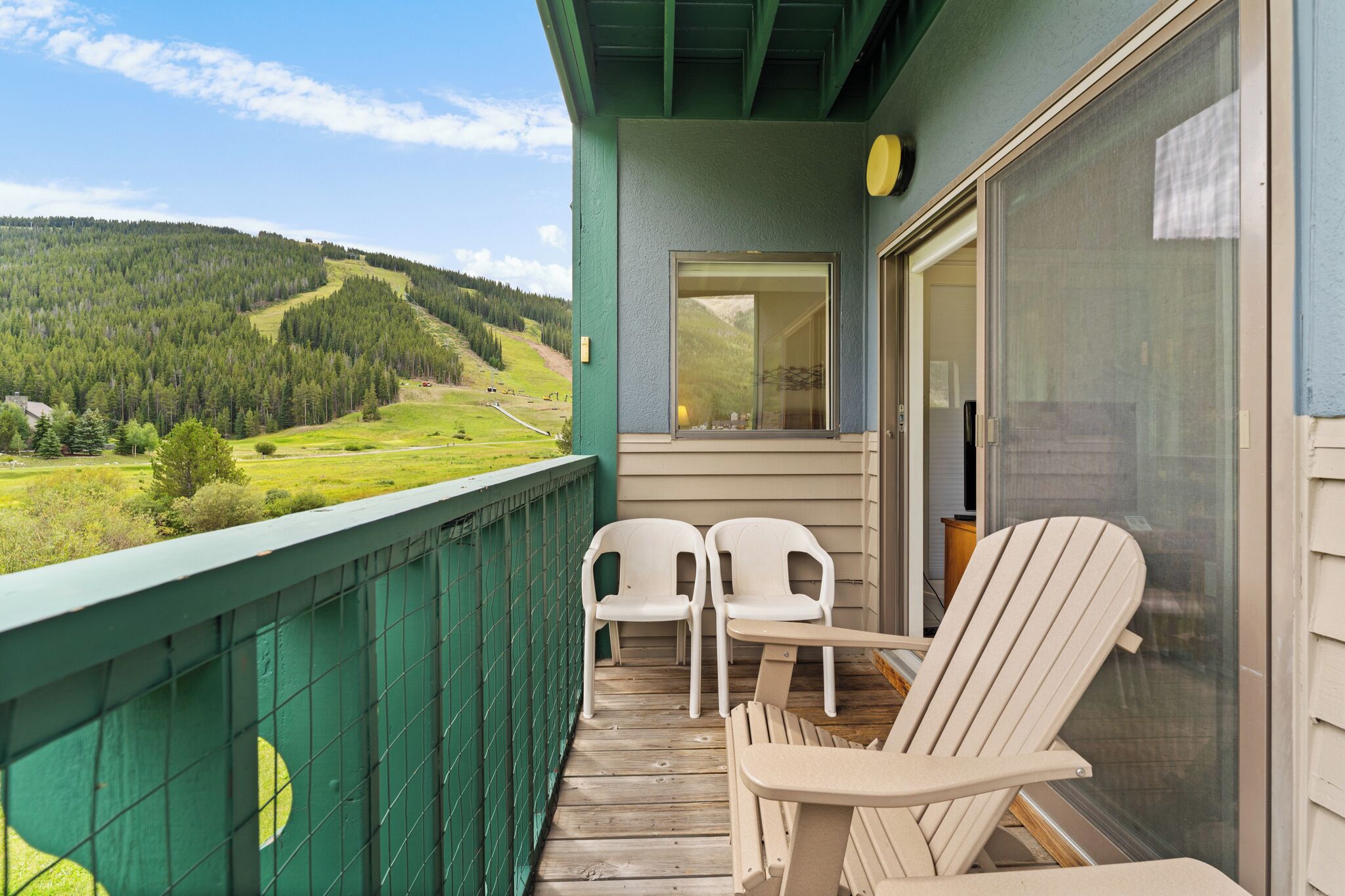 A balcony with two white chairs, a wooden table, and a view of a green landscape with hills.