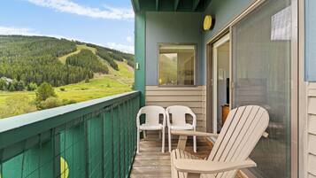 A balcony with two white chairs, a wooden table, and a view of a green landscape with hills.