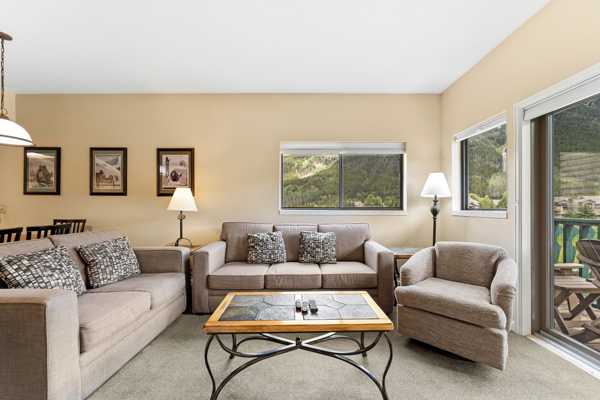 A living room with a beige sofa, a coffee table, and a chair. There are framed pictures on the wall and a window with a view of greenery.