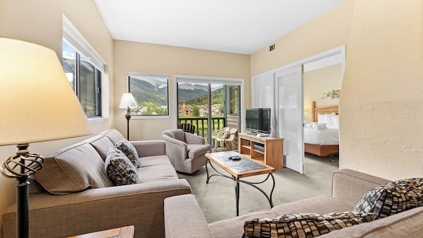 A hotel room with a sofa, armchair, and a small table with a remote control. There is a TV on a wooden stand and a bed with a white headboard. The room has large windows with a view of mountains and houses. - Gac214c1 (Copper Mountain)