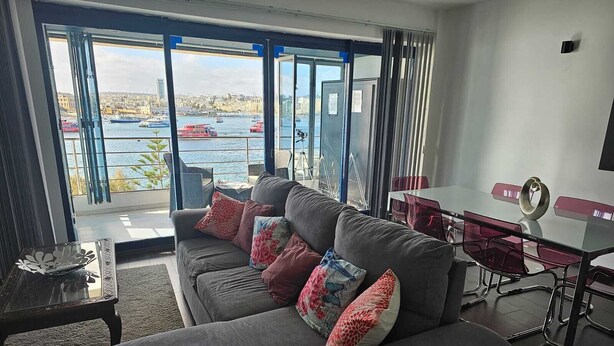 A modern living room with a grey sofa, red and floral cushions, a glass coffee table, and a view of a harbor with boats. - Sea View Modern Apartment in Sliema Ferry 250 sqm (Tas-Sliema)