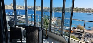 A balcony with wicker chairs and a potted plant, overlooking a harbor with boats and a cityscape.
