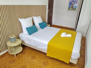 Un lit bien fait, avec des oreillers blancs et bleus, une couverture jaune et une serviette enroulée, posés sur un plancher en bois au motif décoratif.