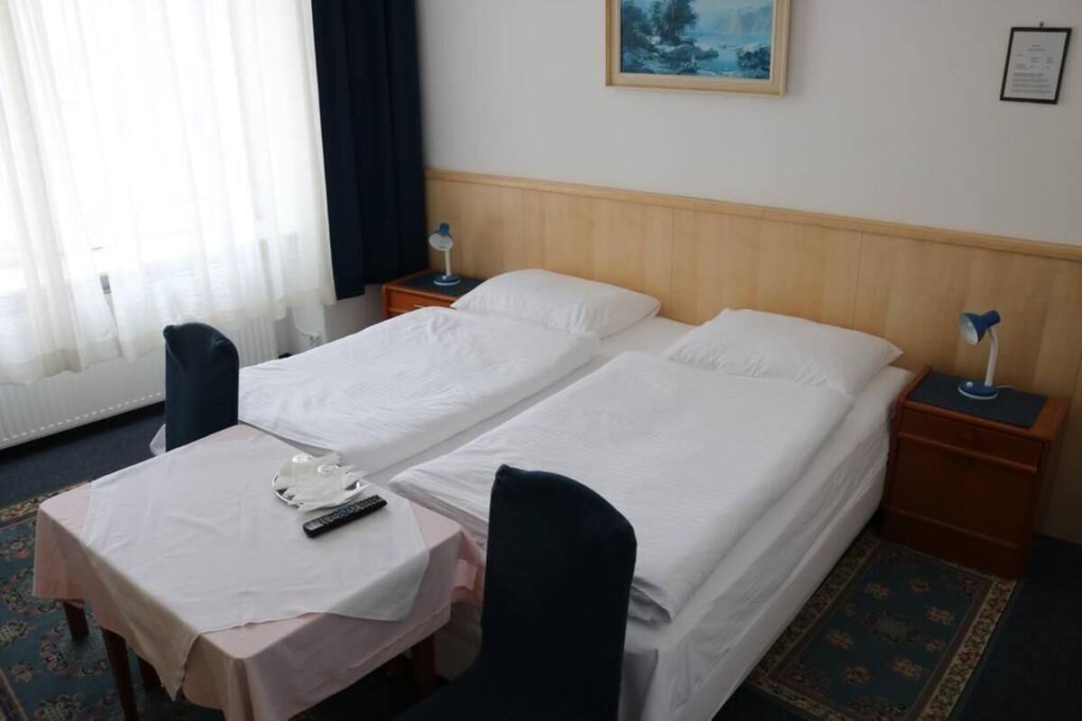 Habitación de hotel con dos camas individuales, una mesita pequeña con una silla, una mesita de noche con lámpara y una ventana con cortinas traslúcidas.