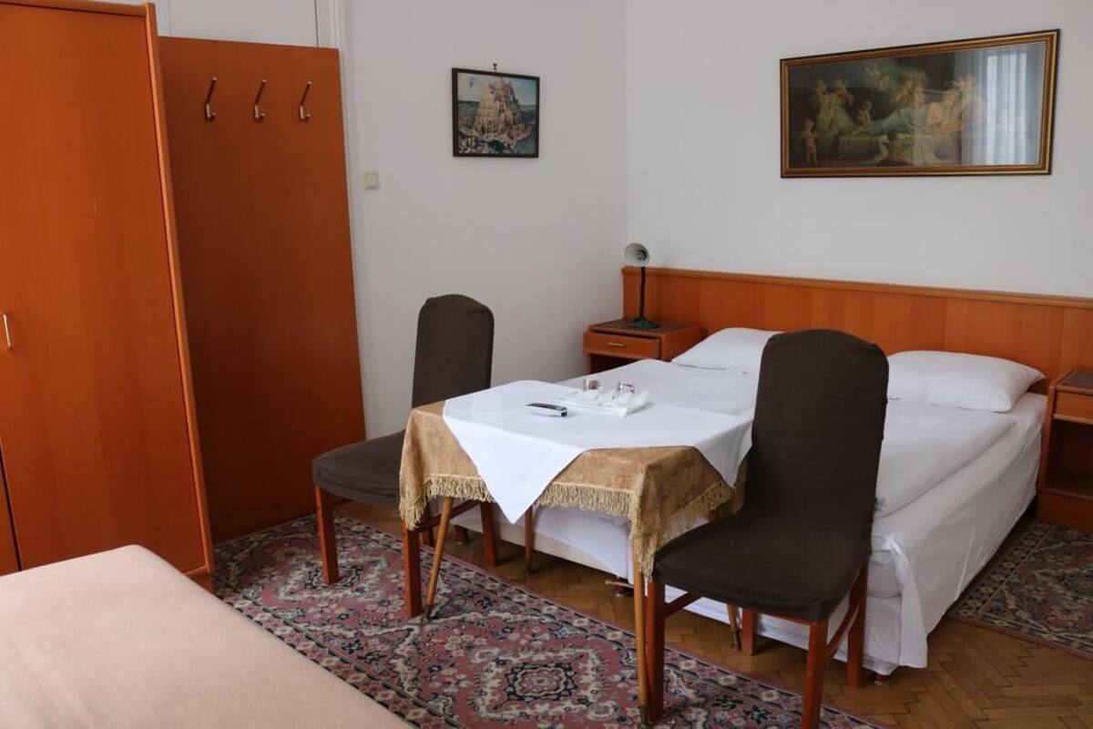 Habitación de hotel con una cama, una mesa con mantel, dos sillas, una mesita de noche, una lámpara y un armario.