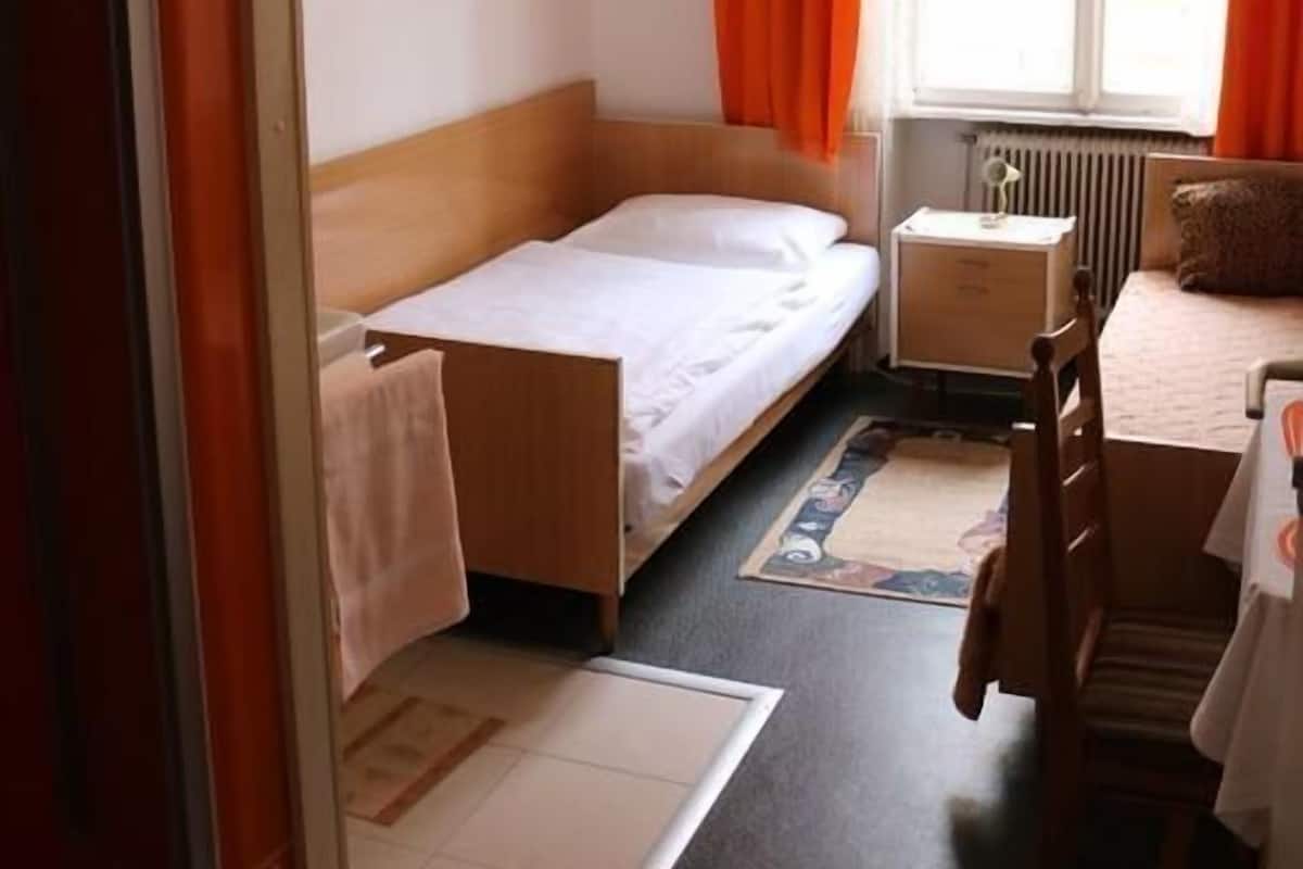 Una habitación pequeña con una cama individual, cabecera de madera, una mesita de noche, una silla y un ventanal con cortinas anaranjadas.