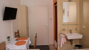 Ein Hotelzimmer mit einem Bett, einem Tisch mit weißer Tischdecke und orangefarbenen Kreisen, einem an der Wand montierten Fernseher, einem Badezimmer mit Waschbecken und Spiegel sowie einer Holztür.