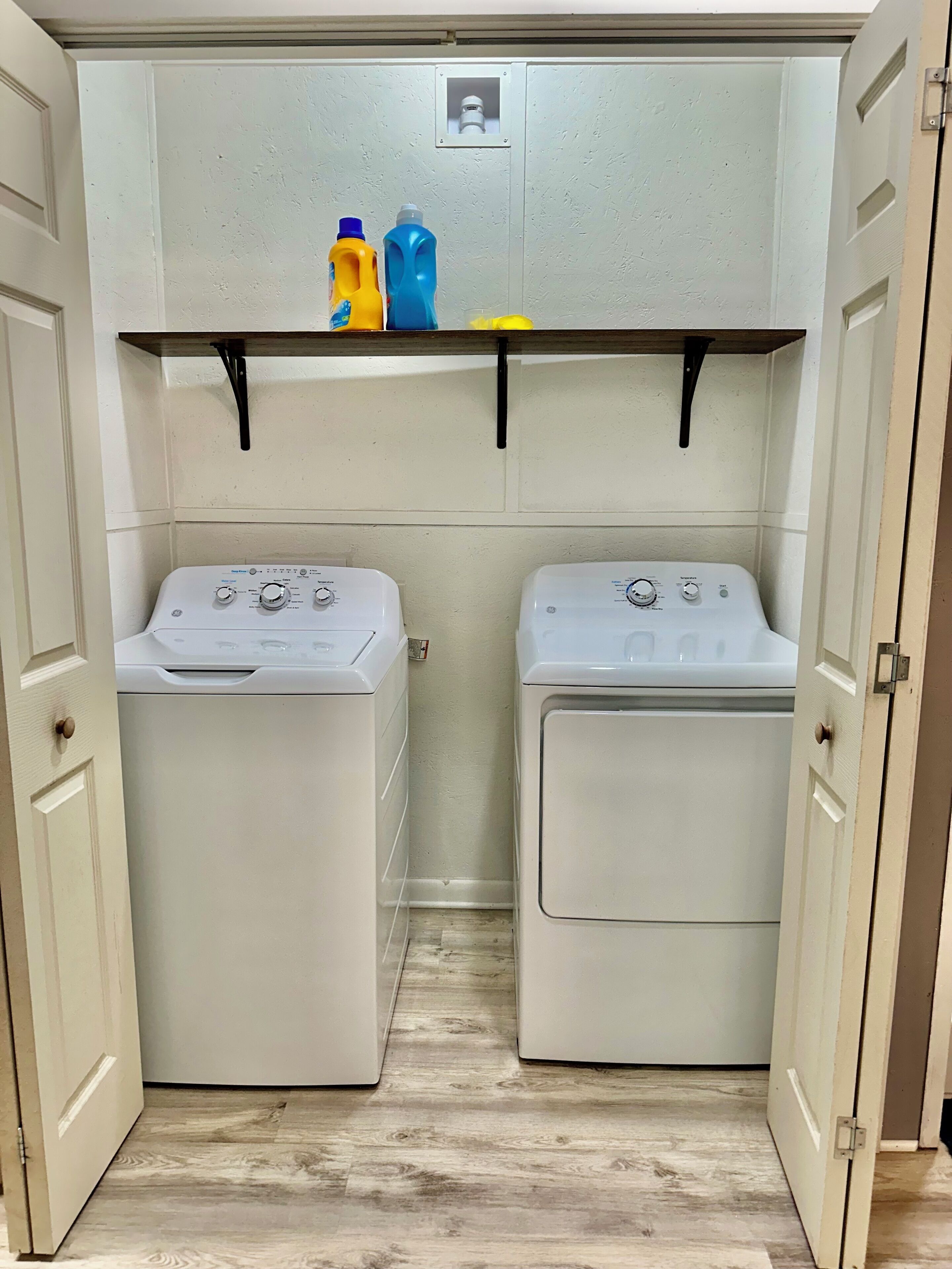 A laundry room with two front-loading washing machines, a shelf with cleaning supplies, and a small window above the machines.