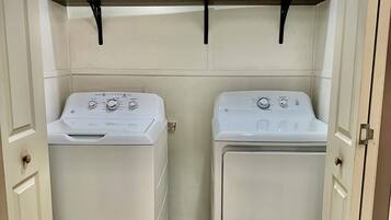 A laundry room with two front-loading washing machines, a shelf with cleaning supplies, and a small window above the machines.