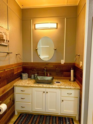 Une salle de bain avec un meuble-lavabo en bois, un miroir rond, un lavabo avec une cuve sur pied et un distributeur de serviettes.