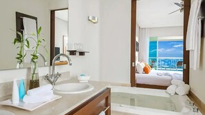 Uma casa de banho moderna com uma banheira grande, uma pia e um espelho. Dá para um varanda com uma cama e vista para o oceano.
