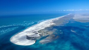 An aerial view of a coastal area with a sandy beach, clear blue waters, and a distant shoreline.