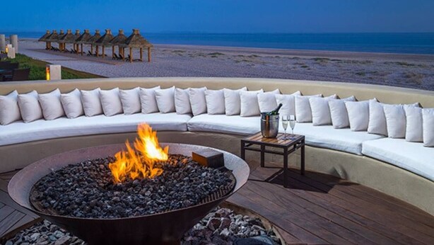 A beachside seating area with a fire pit, a table with drinks, and a view of the ocean.