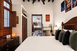 A hotel room with a bed, a desk, a chair, a heater, a ceiling fan, and a kitchen visible through an open door.