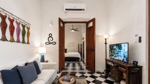 Habitación de hotel con piso a cuadros, un sofá blanco, un televisor y un pasillo que conduce a un dormitorio.