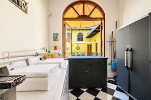 A hotel room with a checkered floor, a bed with white linens, a large black cabinet, and a view of the interior through an arched doorway.