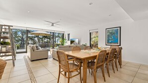 Een ruime eethoek met een grote tafel en zes rieten stoelen, een bank en een televisie in een modern huis met grote ramen en uitzicht op de omgeving.
