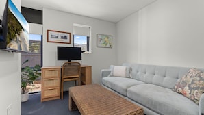 Een compacte leefruimte met een grijze bank, een houten bureau met een computer, een rieten bankje en een aan de muur bevestigde televisie.
