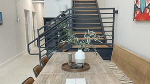 En matplats med ett rustikt träbord, stolar och en vas med gröna växter. I bakgrunden syns en trappa med ett metallräcke.