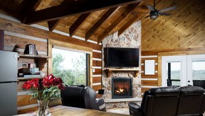 Een gezellig interieur van een chalet met een stenen open haard, houten balken, een flatscreen televisie en een eethoek met een vaas rode rozen.