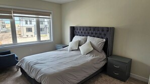 A bedroom with a bed, two bedside tables, a window with a view of buildings, and a carpeted floor. - Beautiful Home backing on Welland Canals STR-289 (Welland)