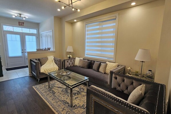 A living room with a sofa, armchairs, a glass coffee table, and a rug. There is a door with an exit sign, a window with blinds, and a lamp on a side table.