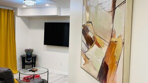 A modern living room with a black leather sofa, a glass coffee table, a mounted flat-screen TV, and a large abstract painting on the wall. - New, Clean and Comfortable Apartment with Private Entrance.  (East Gwillimbury)