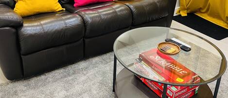 A living room with a black leather sofa, two bright yellow and one pink cushion, a glass coffee table with Monopoly and Scrabble boxes, and a yellow curtain.