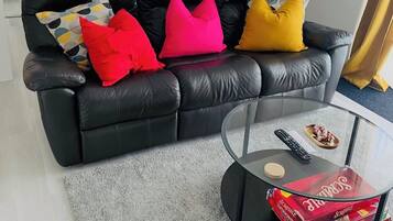 Un salon avec un canapé en cuir noir, une table basse en verre et une boîte de Scrabble posée sur la table.