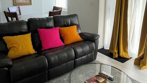 A living room with a black leather sofa, colorful cushions, a glass coffee table with a Monopoly game box, and a dining area in the background. - New, Clean and Comfortable Apartment with Private Entrance.  (East Gwillimbury)