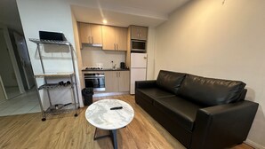 A compact living space with a kitchenette, a black leather sofa, a round coffee table, and a metal shelf unit. - One Bedroom Apartment (Hamilton)