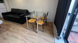 A compact living space with a black leather sofa, a glass-top dining table with four chairs, and a sliding glass door leading to an outdoor area. - One Bedroom Apartment (Hamilton)