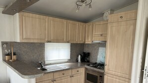 A kitchen with wooden cabinets, a sink, a stove, and a window with blinds. - ZP 468 - Camping de Zandput (Vrouwenpolder)
