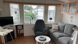 A living room with a grey sofa, a small round table, a grey armchair, a television, and a window with blinds. - ZP 468 - Camping de Zandput (Vrouwenpolder)