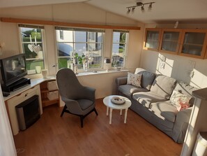 Ein gemütliches Wohnzimmer mit Fernseher, einem grauen Sessel, einer Couch mit floralen Kissen, einem kleinen weißen Tisch und großen Fenstern mit Blick ins Freie.
